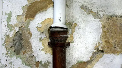Peeling, water-stained wall plaster around a rusted drain pipe