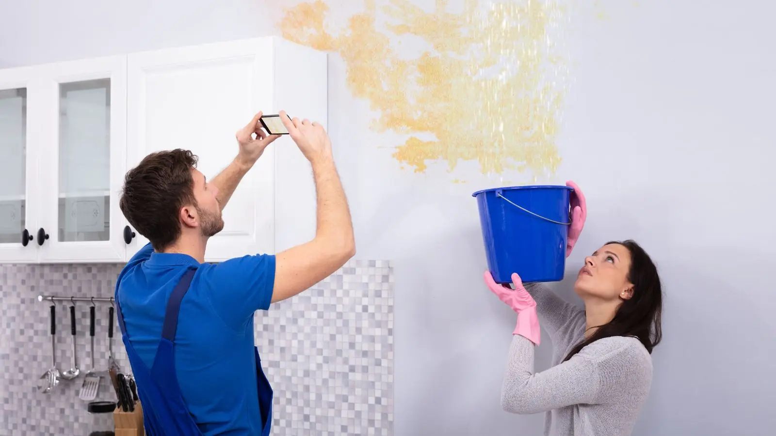 Couple photographing a water stain on a kitchen wall while holding a bucket to document the damage